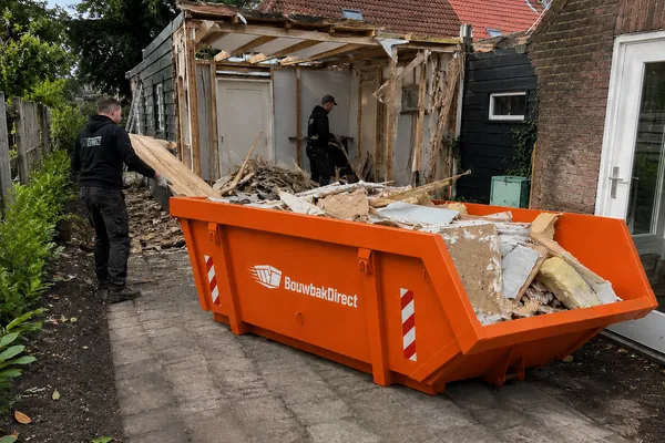 Afvalcontainer naast een schuur die wordt gesloopt, gevuld met hout en plaatmateriaal